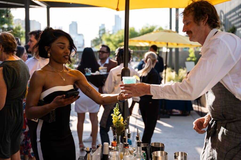 a social gathering on a outside patio, featuring a bartender serving a cocktail to a guest.