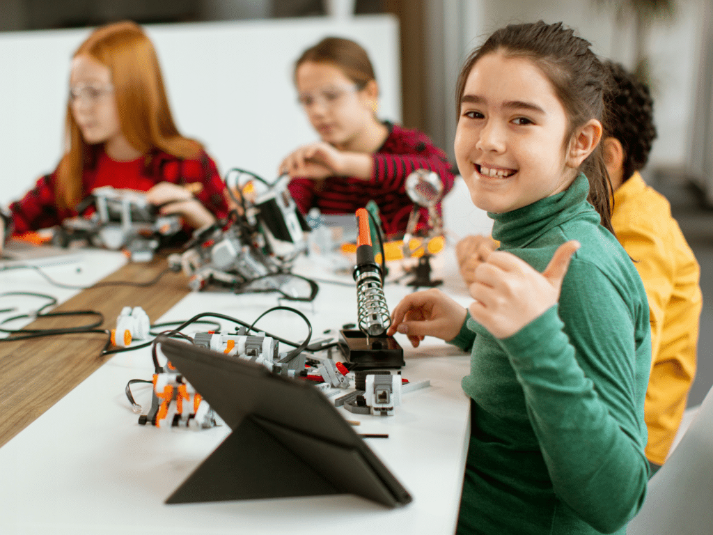 Children sitting at a table engaging in robotics activities. A girl in a green shirt smiles and gives a thumbs-up, showcasing the enthusiasm of girls in STEM. Various electronic components and a tablet are on the table, teaching young entrepreneurs the foundations of technology.