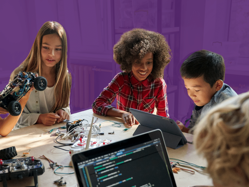 Four children are engaging in STEM activities at a table. One girl is holding a robotic car, another girl and a boy are looking at a tablet, and a laptop with code is visible in the foreground, showcasing diversity in entrepreneurship education.