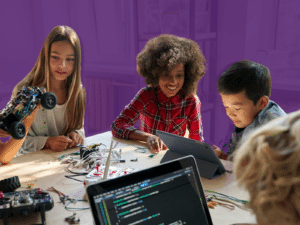 Four children are engaging in STEM activities at a table. One girl is holding a robotic car, another girl and a boy are looking at a tablet, and a laptop with code is visible in the foreground, showcasing diversity in entrepreneurship education.