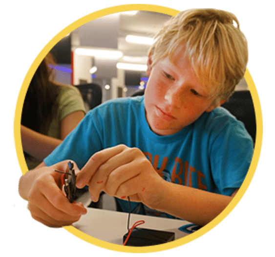 A young person with blonde hair, wearing a blue shirt, is focused on working with electronic components at a table, as part of their k12 entrepreneur curriculum.