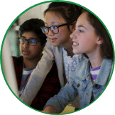 Three young students, two girls and a boy, intently look at a computer screen in their social entrepreneurship program, showing curiosity and engagement. The boy wears glasses.