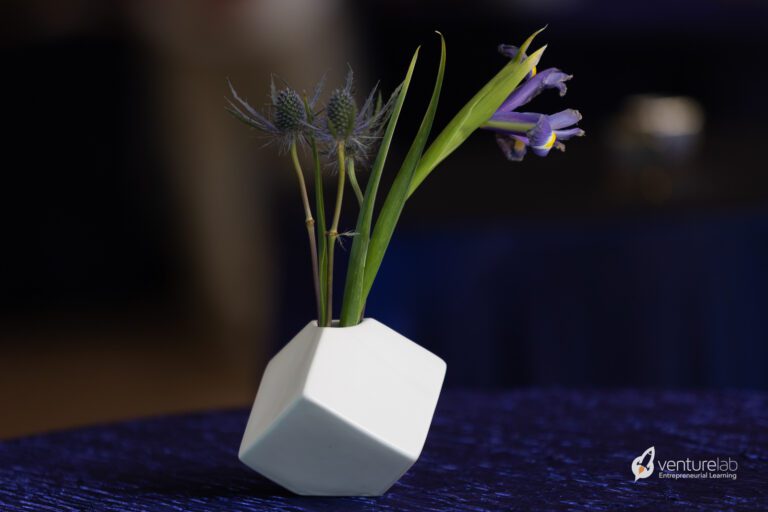 A small white geometric vase holds a purple iris and green foliage, placed on a dark blue surface. The text 
