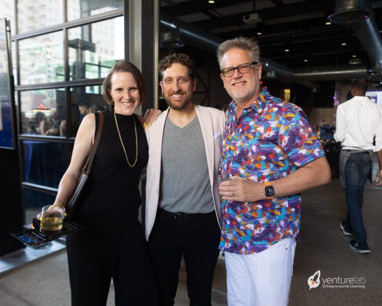 Three people are standing together in an indoor event space, smiling at the camera. One is holding a drink. The person in the middle wears a blazer and grey shirt. The VentureLab logo, known for promoting youth entrepreneurship education, is visible in the corner.