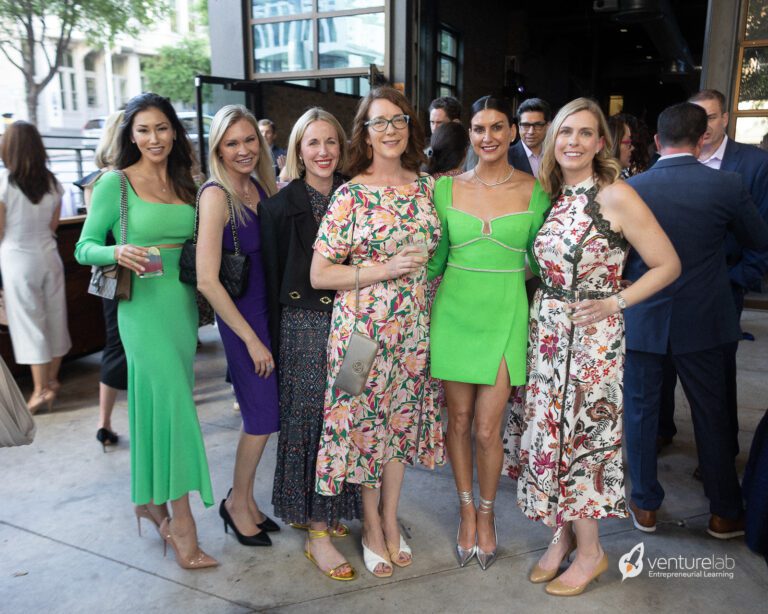 A group of six women poses together at an indoor event, dressed in various bright and floral outfits. People mingle in the background near large open windows. A 