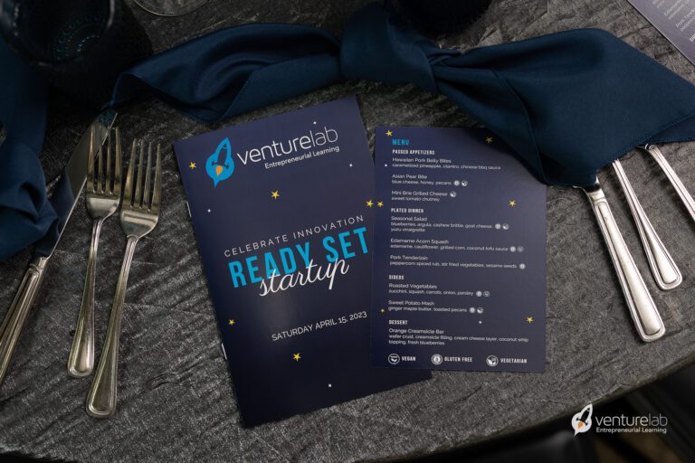 A table setting with silverware and a dark blue napkin surrounds a VentureLab flyer and menu for an event, 