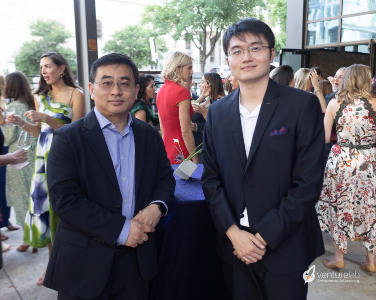 Two men in business attire stand in the foreground at a social event, discussing youth entrepreneurship education, with people mingling in the background near a high-top table.
