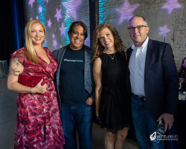 Four people stand together, smiling at an indoor event with a backdrop of a brick wall and starry pattern lighting; an emblem with 