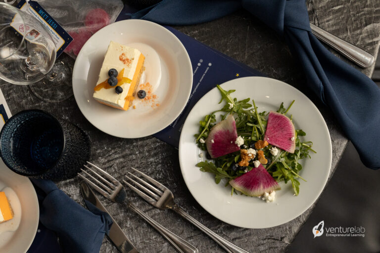 A table setting with a plated salad featuring watermelon radishes and a plate of dessert with blueberries. Napkins are neatly folded, and utensils are arranged beside the plates, reflecting the meticulous attention to detail often emphasized in youth entrepreneurship education. The VentureLab logo is visible.