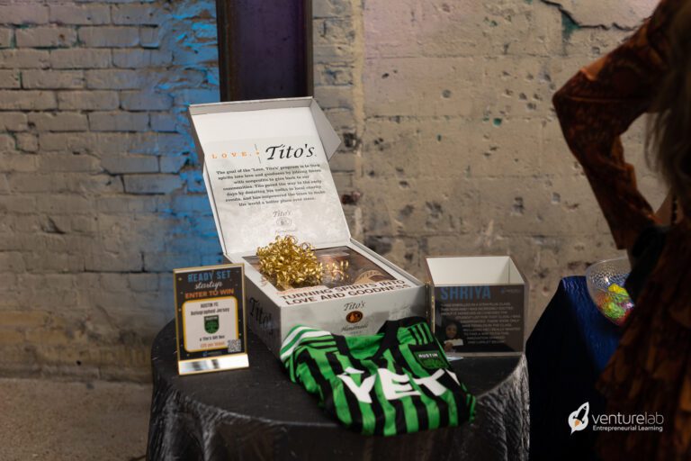 A display table featuring an open gift box with a golden ribbon, a green and black striped shirt, a sign, and other materials highlights youth entrepreneurship education. A brick wall in the background and a 