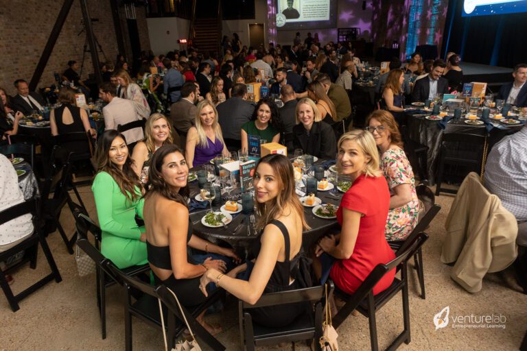 A large group of people seated around tables at a formal event, with some individuals posing for a photo. The venue is well-lit and decorated, and a presentation on youth entrepreneurship education is being projected in the background.
