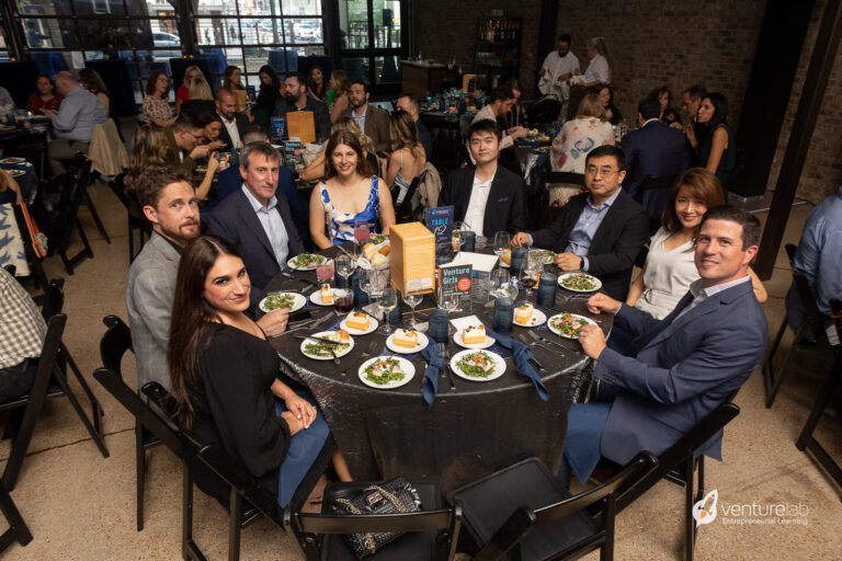 A group of people in business attire are seated around a table at a formal event, enjoying salads and discussing the benefits of entrepreneurship education. Others are seen interacting in the background, highlighting their experiences with teaching entrepreneurship.