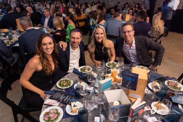 A group of four people seated at a table with salads, desserts, drinks, and event materials during a formal gathering focused on youth entrepreneurship education. Others are seen seated and engaging in conversation in the background.