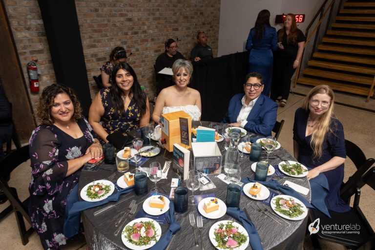 A group of five people sits at a round table with place settings, salads, and small cakes. Behind them are a brick wall, a staircase, and more attendees. The event features a VentureLab banner promoting youth entrepreneurship education.