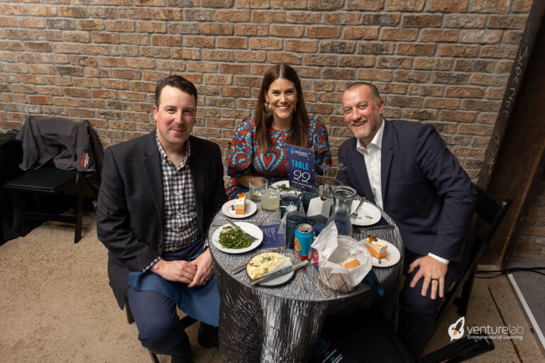Three individuals smiling and seated at a small round table, enjoying a meal. The table has plates of food and drinks. A VentureLab sign is visible, highlighting their commitment to youth entrepreneurship education. The background features a brick wall.