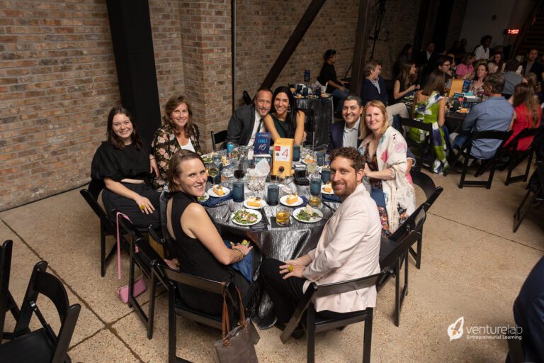 A group of nine people sit around a round table with meals and drinks in a large, indoor venue with brick walls, discussing youth entrepreneurship education. A sign with the number 4 is placed at the center of the table.