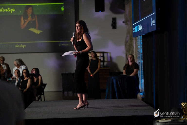 A woman in a black dress is holding a microphone and speaking on stage at an event about teaching entrepreneurship, with a projected image of herself behind her. The event, focused on entrepreneurship education, has several seated attendees.