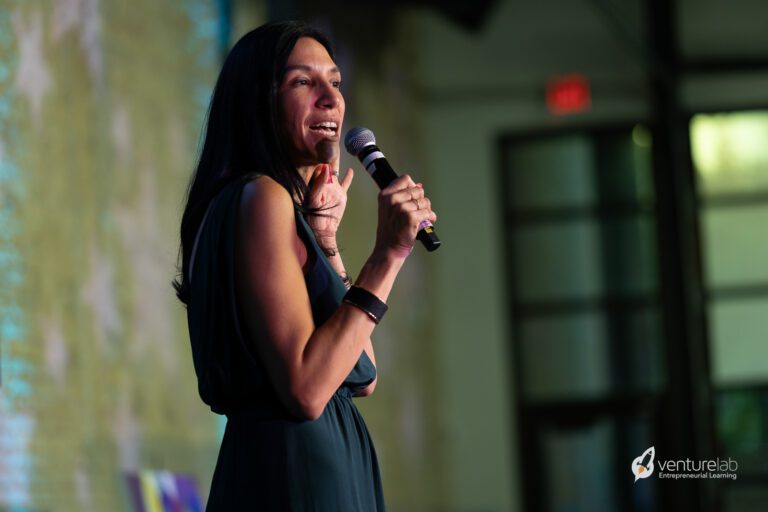 A woman speaks into a microphone while standing on a stage, exuding confidence in her dark sleeveless dress. The blurred background enhances the focus on her, as she discusses youth entrepreneurship education. The VentureLab logo is visible in the bottom right corner, underscoring the event's theme.