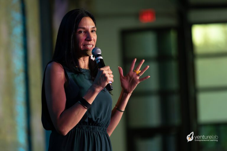 A woman in a dark sleeveless dress speaks into a microphone on a stage. The background is softly lit and out of focus, and the VentureLab logo is visible in the bottom right corner, highlighting their commitment to youth entrepreneurship education.