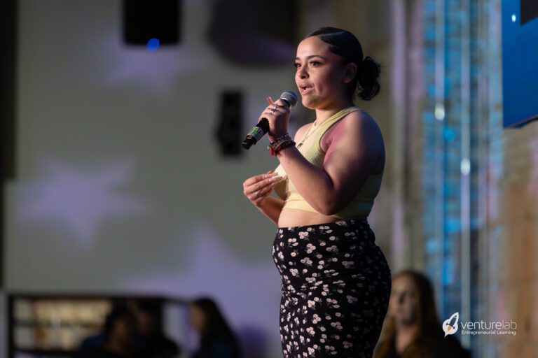 A person stands on stage holding a microphone, wearing a yellow top and patterned skirt. The background shows blurred audience and a 