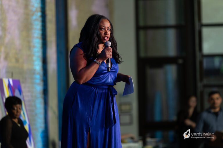 A woman in a blue dress speaks into a microphone onstage at an event focused on teaching entrepreneurship, with a blurred audience and event backdrop in the background.