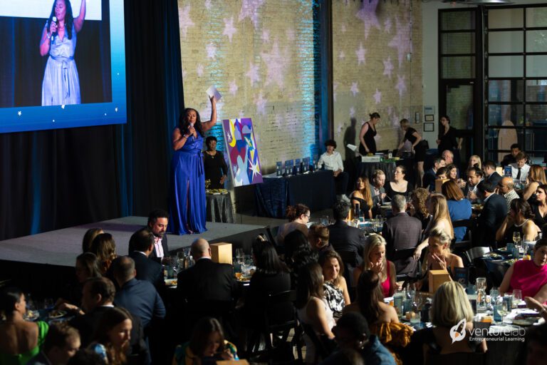 A woman in a blue dress speaks at a podium during the Ready Set Startup Gala, with guests seated at tables and a large screen displaying her image in the background.