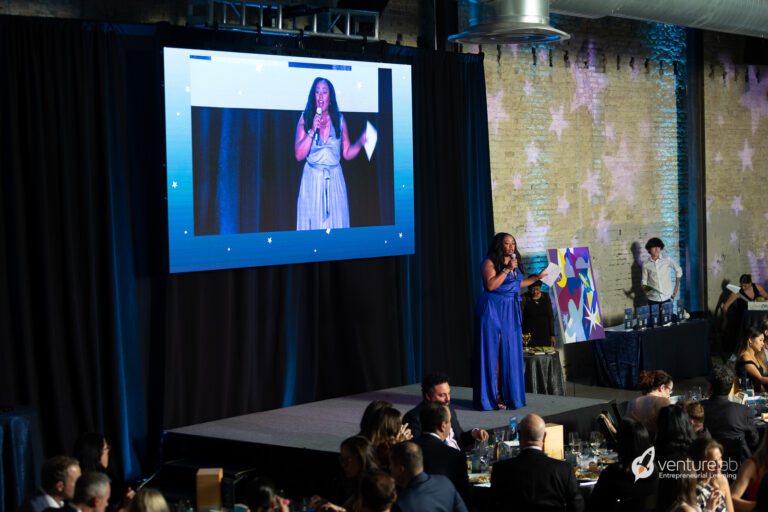 A woman in a blue dress speaks into a microphone on stage with a large screen behind her displaying her image. As she discusses the new youth entrepreneurship education program, an audience at round tables attentively listens.