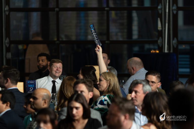 A woman raises a bid paddle at an auction event, surrounded by other seated participants, representing a local nonprofit focused on youth entrepreneurship education.