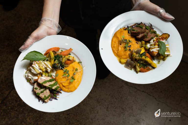 Two white plates of gourmet food are held up by a person wearing gloves. Each plate contains a variety of ingredients, including meat, vegetables, and a bright orange purée—showcasing the creativity one might inspire through youth entrepreneurship education.