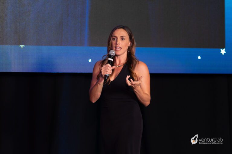 A woman in a black dress speaks into a microphone while gesturing with her left hand on a stage. The background features a large screen and the VentureLab logo in the bottom right corner, reflecting their commitment to entrepreneurship education.
