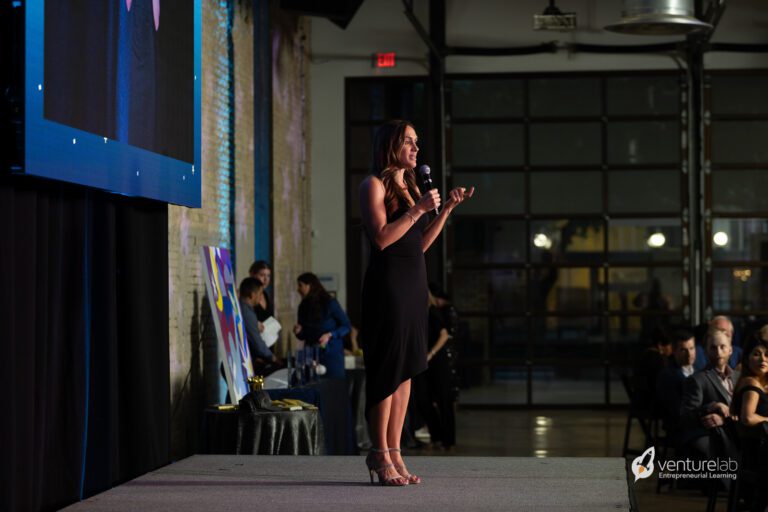 A woman in a black dress speaks into a microphone on stage at a formal event focused on youth entrepreneurship education. A large screen and VentureLab branding are visible in the background. Audience members are seated and watching her intently.