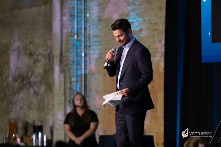 A man in a suit holds a microphone and papers while speaking on a stage, discussing the entrepreneurship education program. A woman stands in the background. The VentureLab logo is visible on the screen behind him.