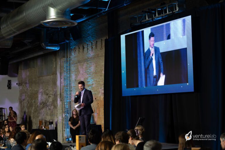 A person speaks into a microphone on a stage at an event, teaching entrepreneurship as their image is projected on a large screen behind them. An audience is seated in front of the stage. The venue has industrial decor.