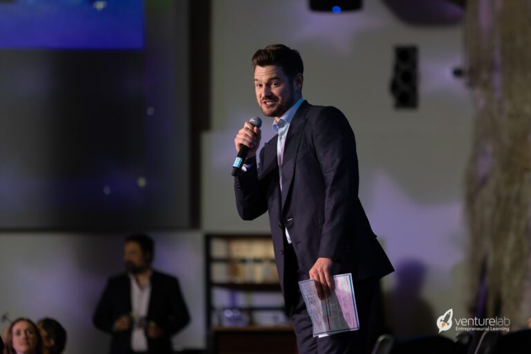 A man in a suit holds a microphone and a document while speaking on stage at an event about teaching entrepreneurship. The VentureLab logo is visible in the bottom-right corner.