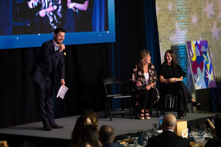 A man speaks into a microphone while two women sit on a stage with a painting in the background, discussing youth entrepreneurship education. An audience is seated in front.