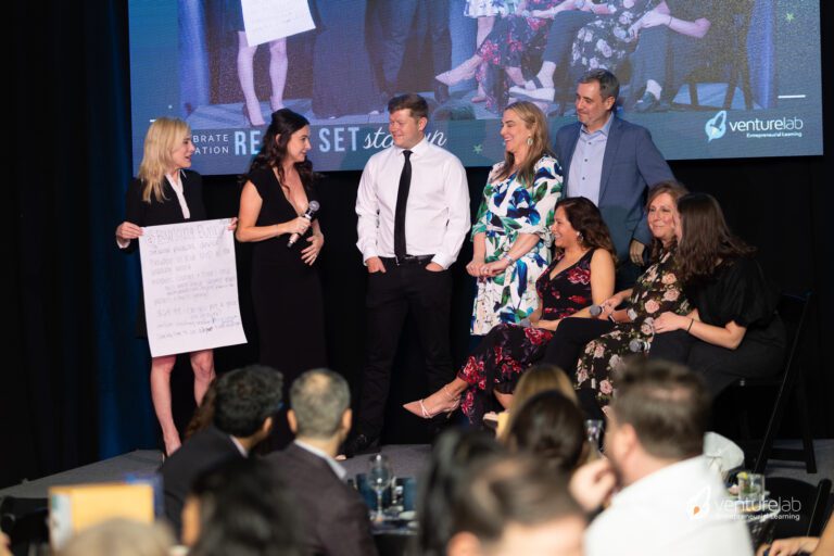 A woman presents a large sheet of paper on stage to a group of four people standing and four people seated, discussing concepts within entrepreneurship education. A screen in the background shows the same scene, with a 