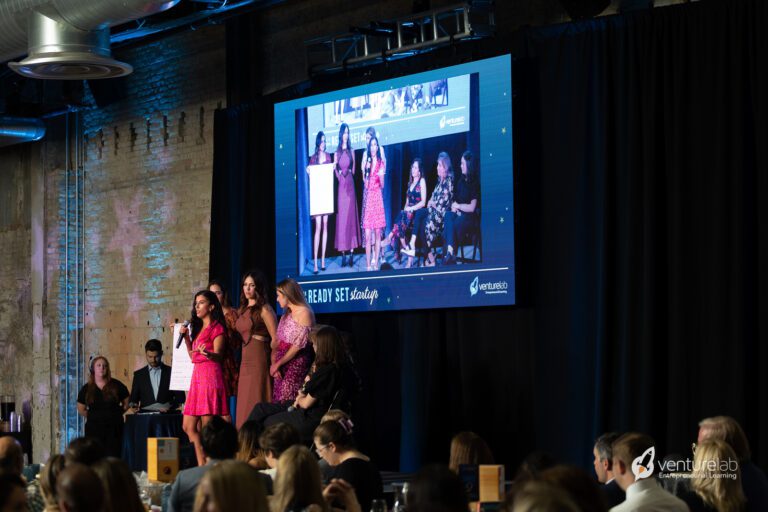 A group of women in pink dresses are speaking on stage in front of an audience. A large screen behind them displays their image. The event, hosted by VentureLab, highlights the importance of youth entrepreneurship education.