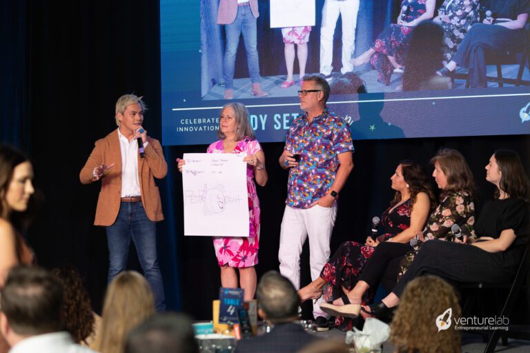 A group of people are on a stage presenting. A man holds a microphone while a woman displays a large sheet of paper with a drawing. Three other people are seated to the right, with a large screen behind them showcasing their youth entrepreneurship education program.