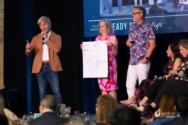 A person in a blazer speaks into a microphone while another holds a large paper with sketches. Others, including a man with a mic, are seated and watching attentively. The setting appears to be an event focused on youth entrepreneurship education.