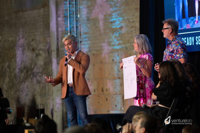 A man with a microphone speaks on stage while two people beside him hold and look at a large piece of paper, presenting to an audience seated in the foreground. The event highlights youth entrepreneurship education, inspiring the next generation of innovators.