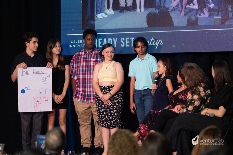 Several young people stand on a stage. One holds a sign showing their project design, showcasing their skills from the youth entrepreneurship education program. Another speaks into a microphone. Two seated adults appear to be listening attentively to the presentation.