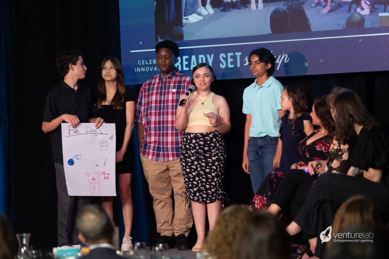 A group of six teenagers stands on a stage. One holds a poster titled 