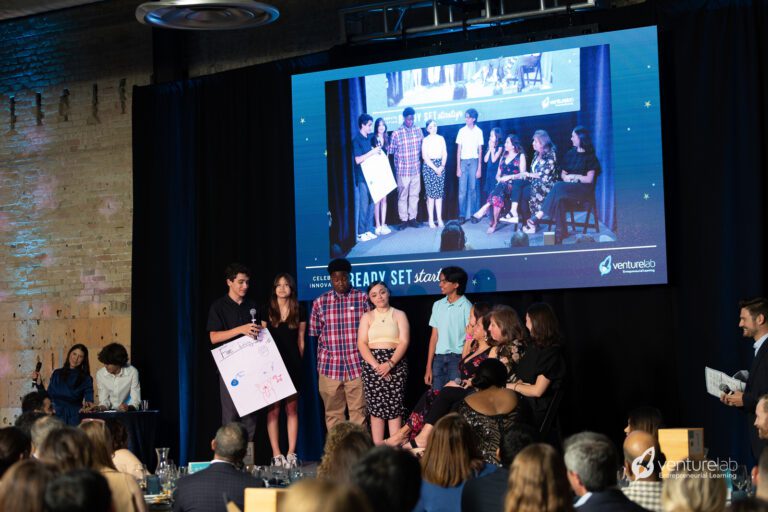 A group of people are presenting on a stage with a large screen behind them displaying their image. The audience seated at tables observes the presentation. The event, hosted by VentureLab, focuses on youth entrepreneurship education.