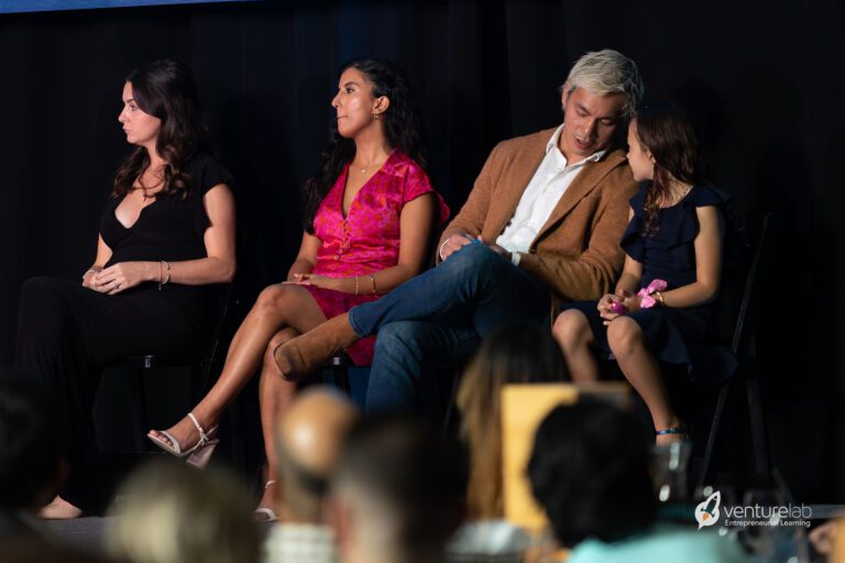 Four people are seated on a stage, two women, one man, and a young girl. The man and girl are interacting, while the two women look forward. 
