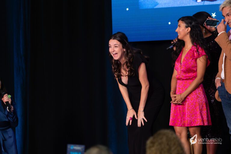 Woman in black dress laughing on stage with a microphone, standing beside another woman in a pink dress. Other people are also present on stage. The 