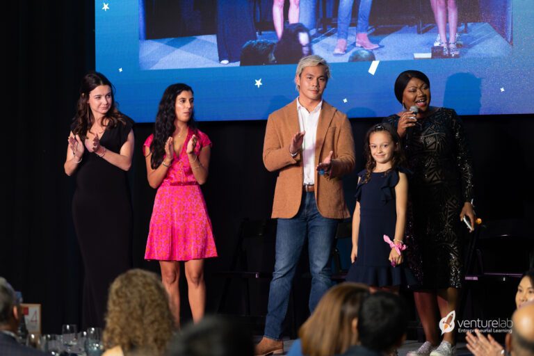 Five people are standing on a stage; four are adults and one is a child. One person is speaking into a microphone about youth entrepreneurship education while the others clap. The background features a large screen displaying an image, highlighting the impact of k12 entrepreneur curriculum.