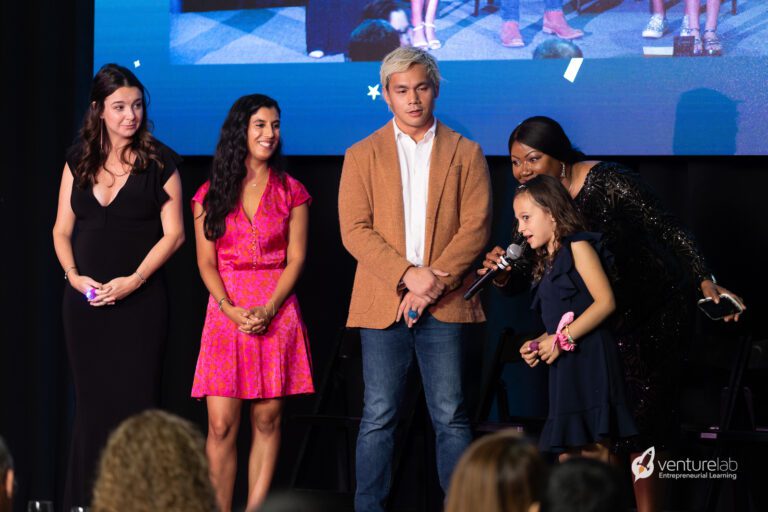 A young girl speaks into a microphone while four adults stand beside her on stage, with one person leaning over her shoulder. A large screen and audience are visible in the background. The logo reads 