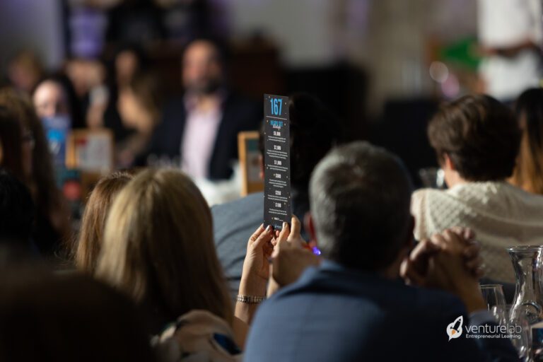 A person holds up a sign with the number 167 at an event, with a crowd watching. The sign has text and numbers, and the logo 