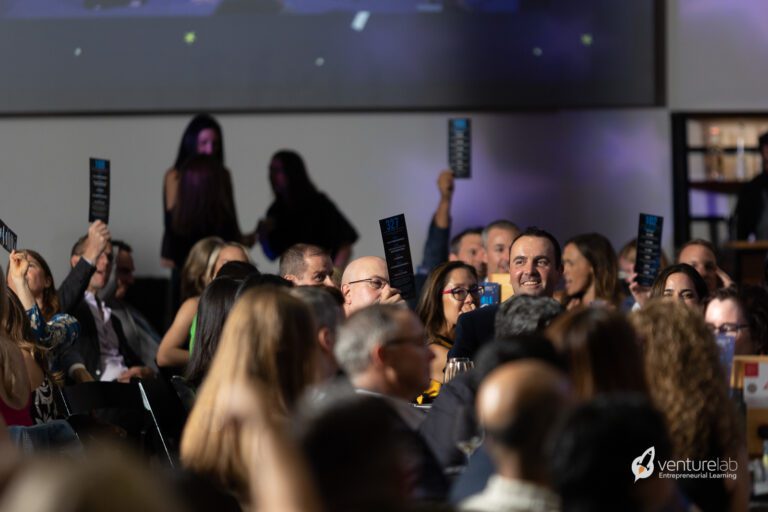 A crowd of people seated at a formal event, many holding up cards or programs. An illuminated screen featuring the VentureLab logo highlights their commitment to teaching entrepreneurship in an engaging and dynamic setting.