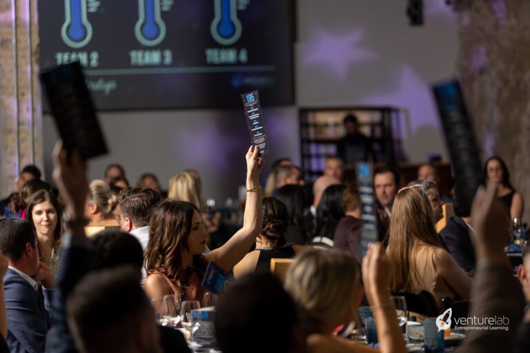 People seated at tables during a formal event, some raising cards. A screen in the background shows team names and scores, highlighting the impact of youth entrepreneurship education on their skills.